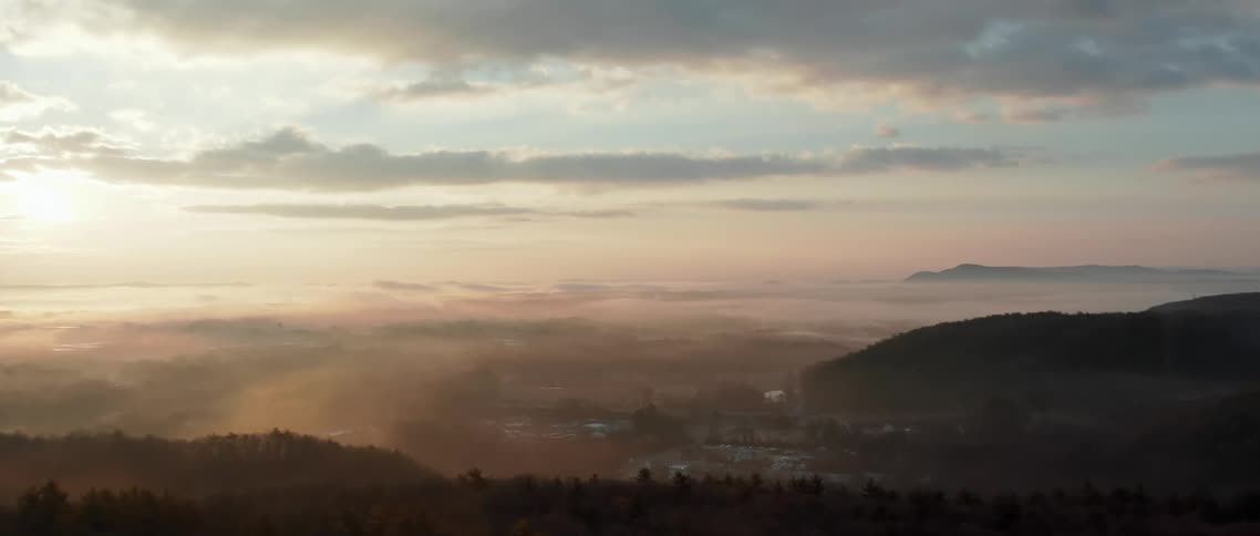 Sun rises over fog filled valley and town surrounded by rounded mountains