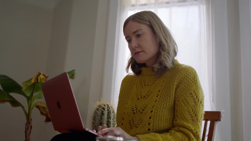 A blonde woman in a yellow sweater uses a laptop in front of a sunny window