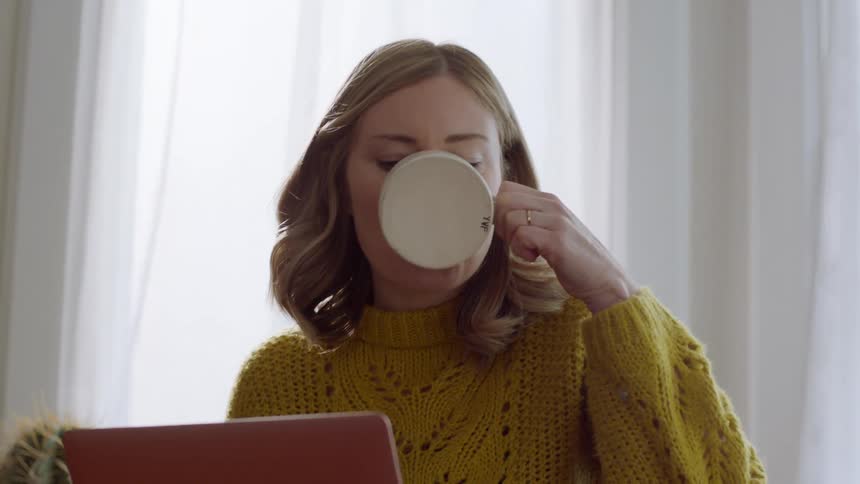 Female sitting on chair in front of a window sipping on a cup of coffee while looking at the screen of a laptop computer that is sitting on the lap of her crossed legs