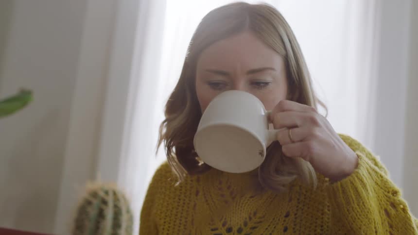 A woman sits at home and drinks out of a mug while using her laptop