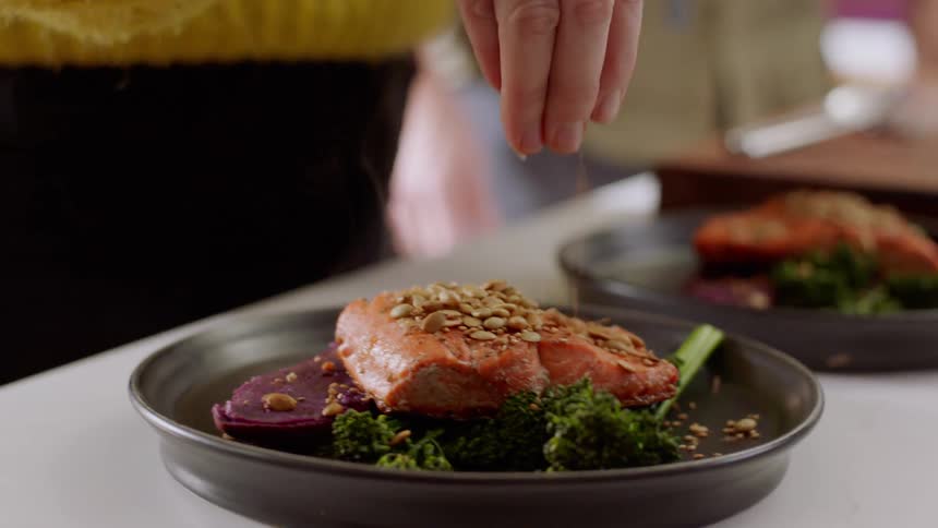 A woman sprinkles some garnish on top of plated salmon and broccoli