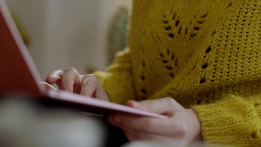 A woman in a yellow sweater sits and uses her pink laptop