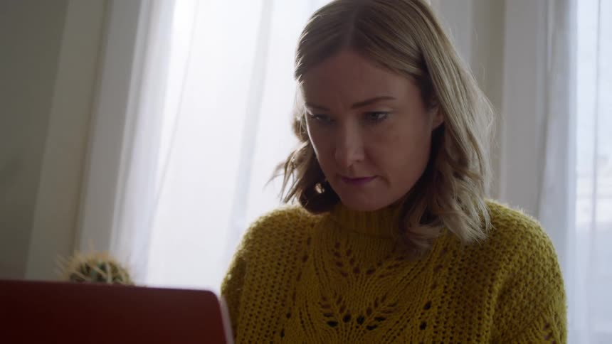 A woman sits with her laptop in her lap typing away with a steaming cup on the table beside her