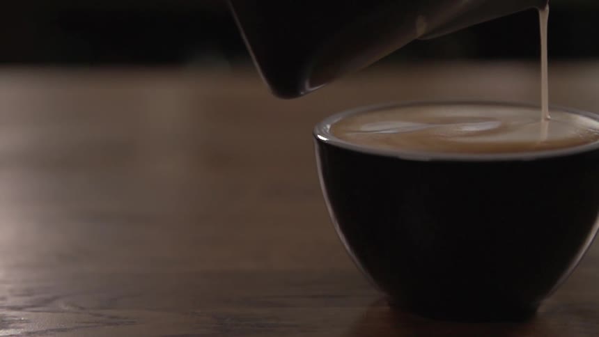 Milk pours from a pitcher into a cup of brown beverage