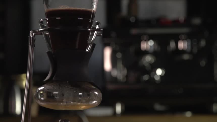 Coffee brews in an old fashioned glass beaker in a coffee house with a large espresso machine in the background