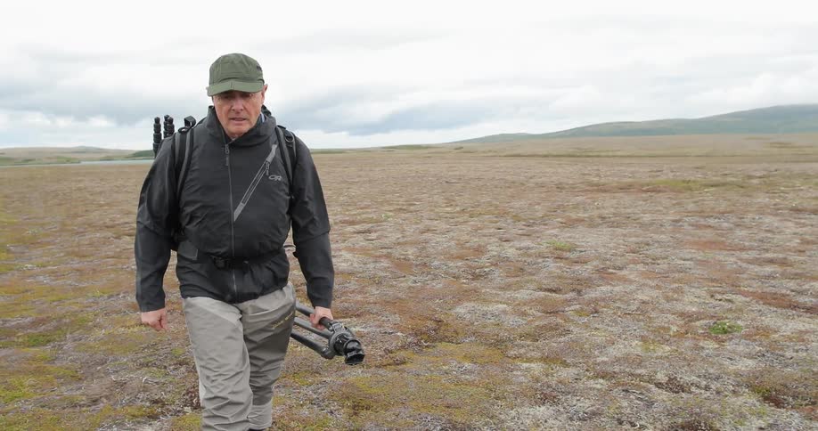 A photographer walks through a field in the wilderness with a tripod in hand