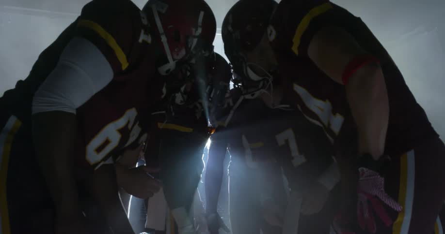 Football players huddle then rise clapping back lit by bright spotlight in fog