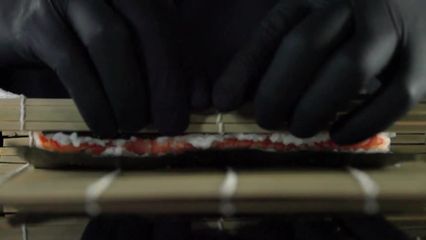 A person wearing black gloves wraps a piece of food in bamboo