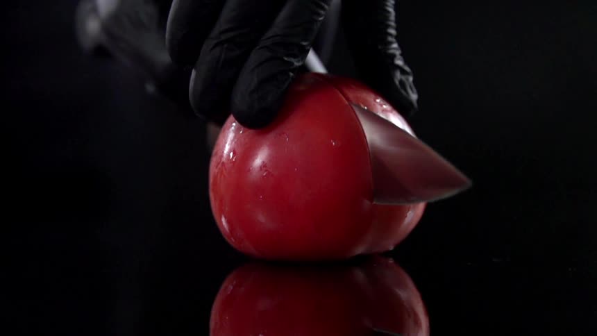 Gloved hands hold and slice a red tomato in half on a black reflective surface