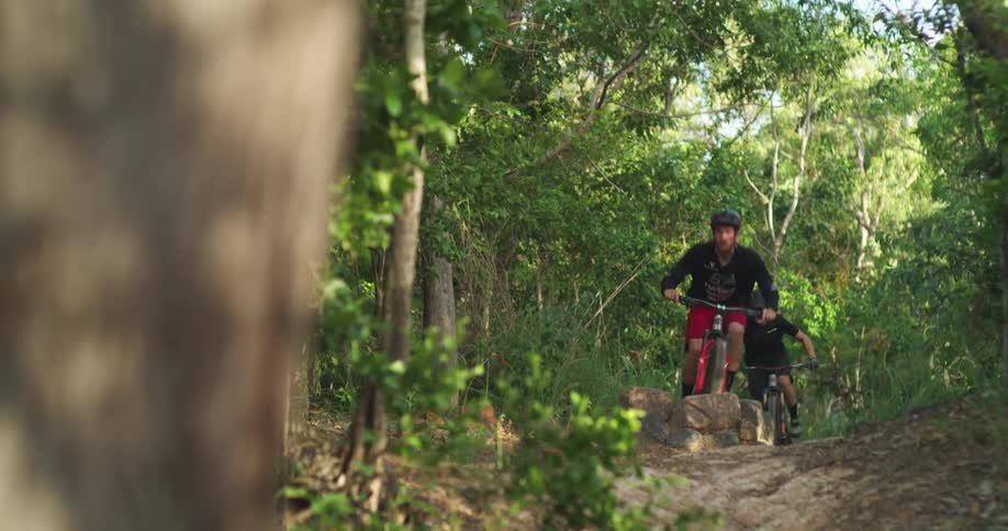 Two bicyclists jump their bikes over a jump in the forest