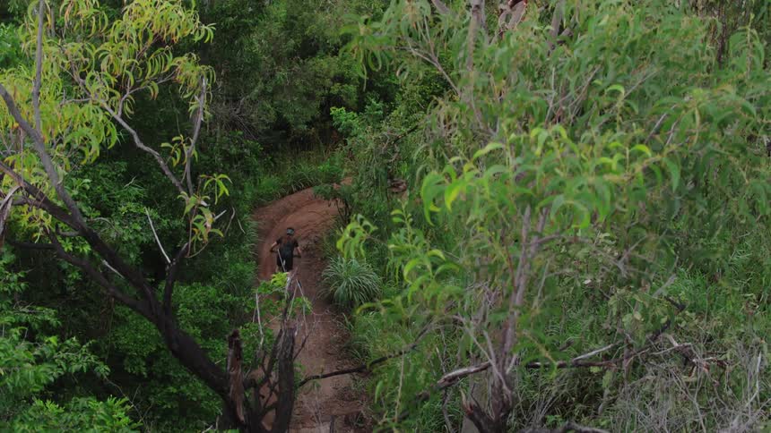 A pair of people mountain bike racing down a narrow dirt path through a forest