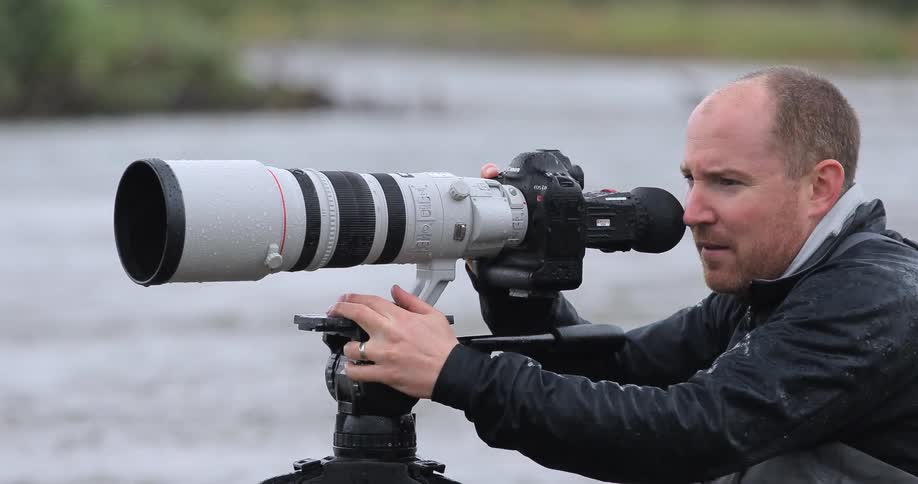 A photographer with a large zoom lens stands near a river