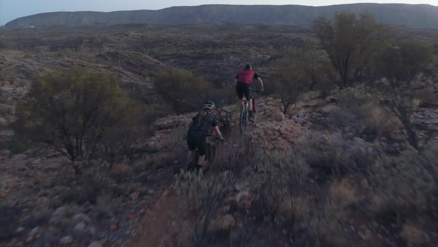 Two people ride mountain bikes down a dirt trail surrounded by brush and rocks