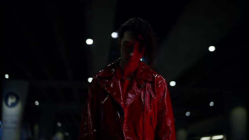 Light reflected from glossy red coat tints womans face red as she paces back and forth in parking garage