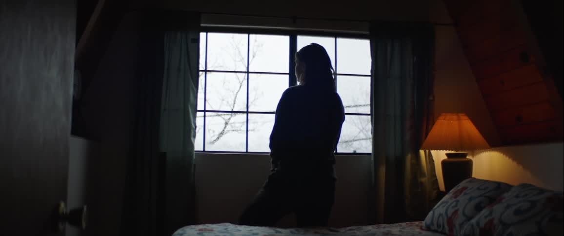A silhouetted woman sits in a dark bedroom while looking out the window