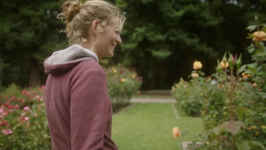 A woman walks through a field with flowers and laughs and stops and smells a rose