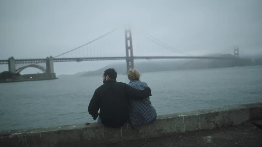 Man sits with arm around woman on concrete ledge overlooking golden gate bridge