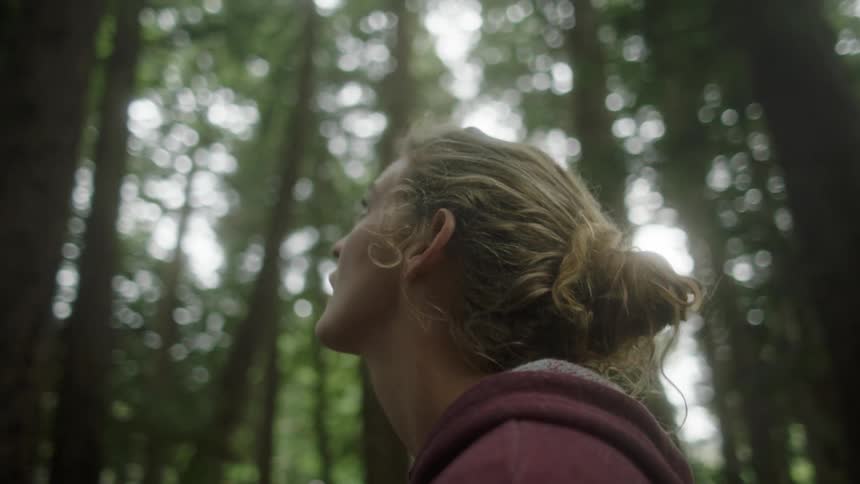 A white woman stands in the woods and looks up at the sky