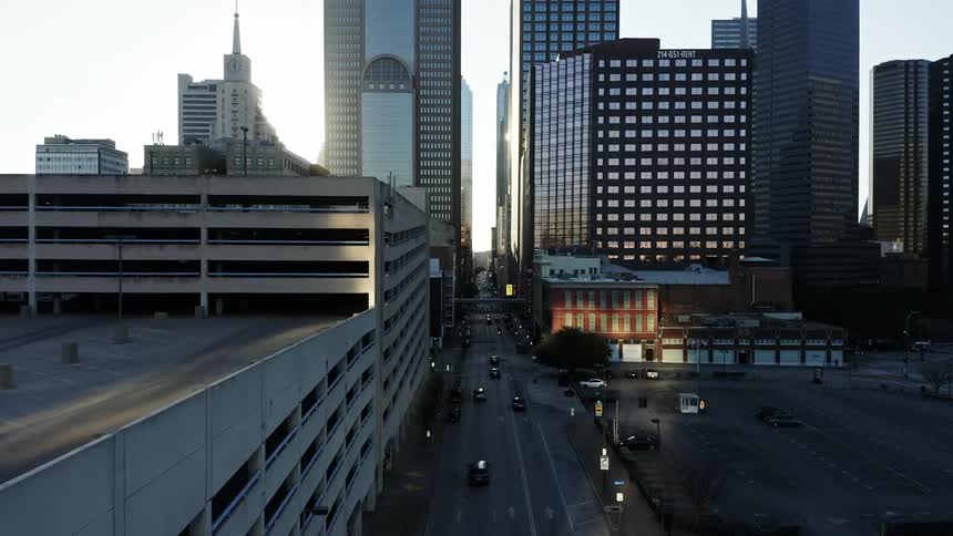 A city street with vehicles next to parking lots with buildings in the background
