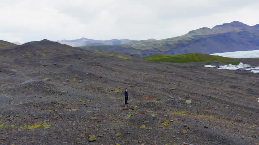 A man wearing a suit stands in a rocky area near some mountains