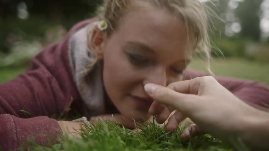 A woman lies in the grass as a man strokes her hair and nose