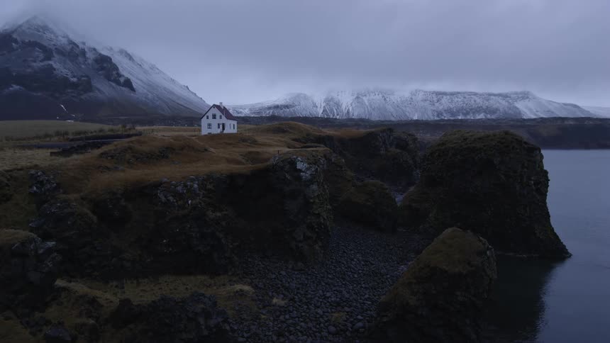 Lone white house near rugged coastline in front of snowy mountain range