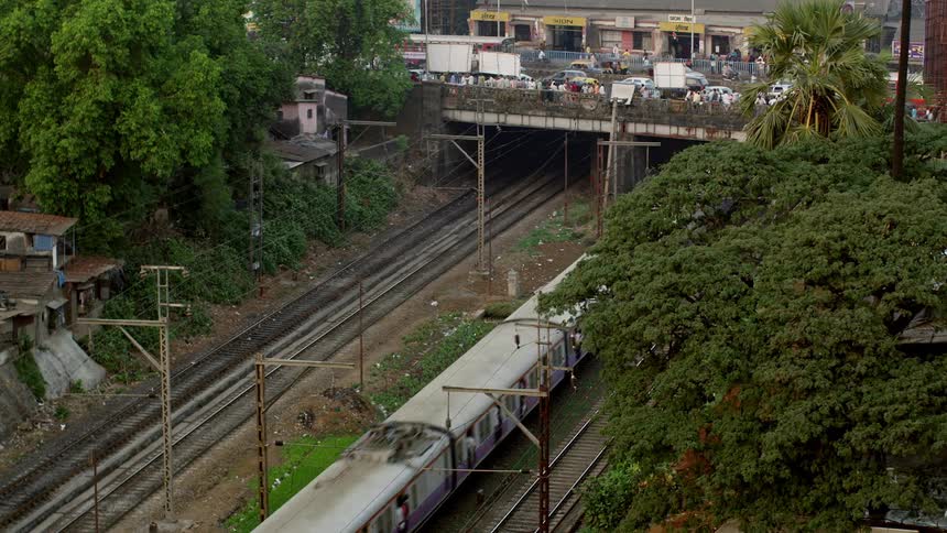 A train travel on rail under a bridge while pedestrians and traffic moves over the top of it