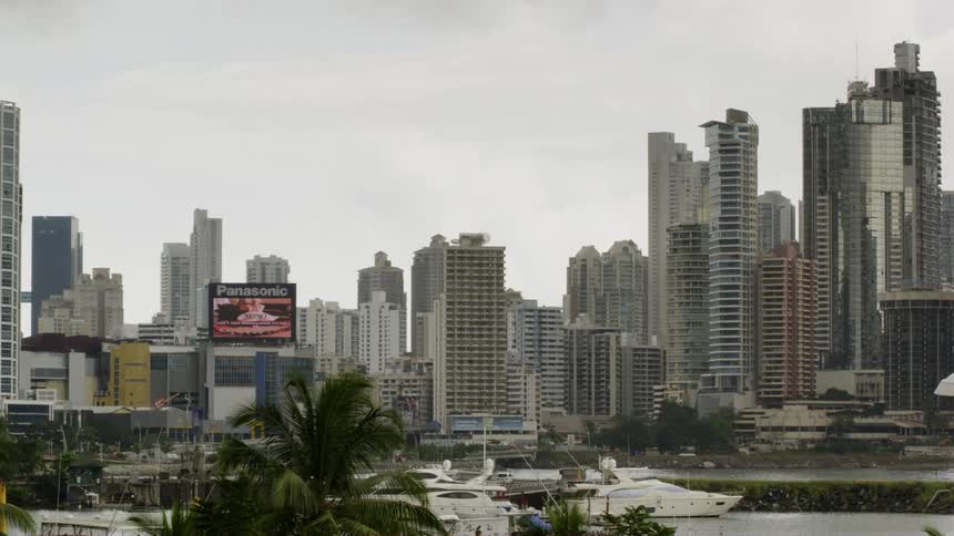 An electronic billboard flashes video above yachts and boats docked in a marina with city skyline behind
