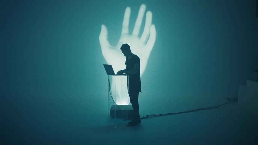A man sways to music as he uses a laptop with a large white hand projected on the wall behind him