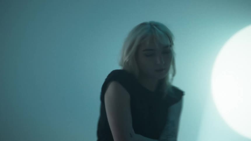 Blonde woman paces back and forth near spotlight projected onto wall