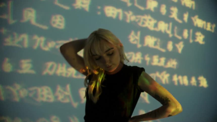 Blonde woman changes poses in from of text projected onto wall