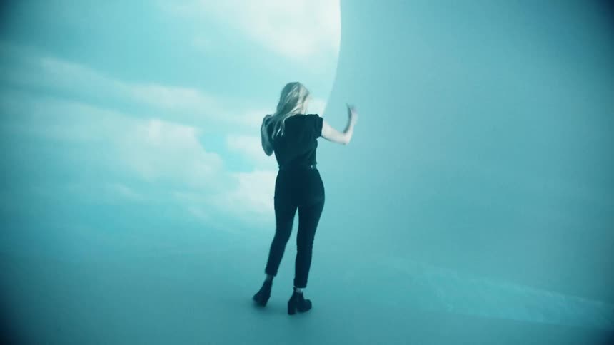 A blonde white woman in black clothing moves around in front of a large projection screen