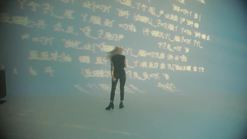 A white woman in black clothing walks around in front of a giant screen with text flashing on it