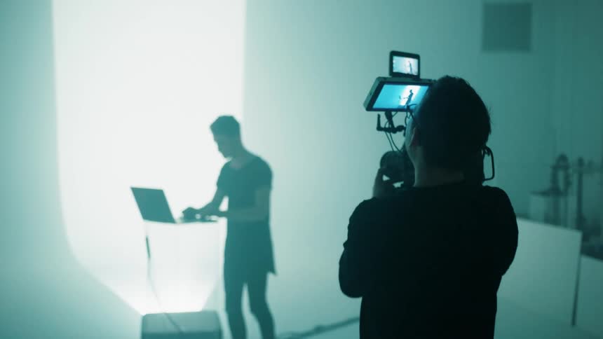 A man with a professional camera walks to another man with a laptop in a studio