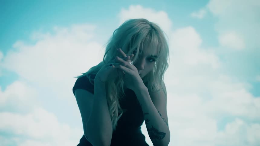 A blonde woman poses in front of a green screen with a sky digitally shown on it
