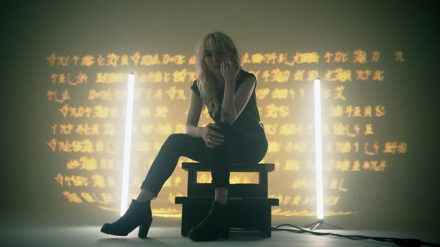 Woman sits near vertical mounted fluorescent light bulbs near wall with text projection
