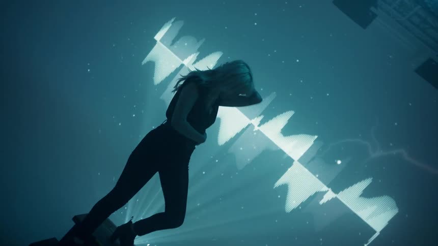 A blonde woman poses on a stand in front of a wall where images and lights are flashing