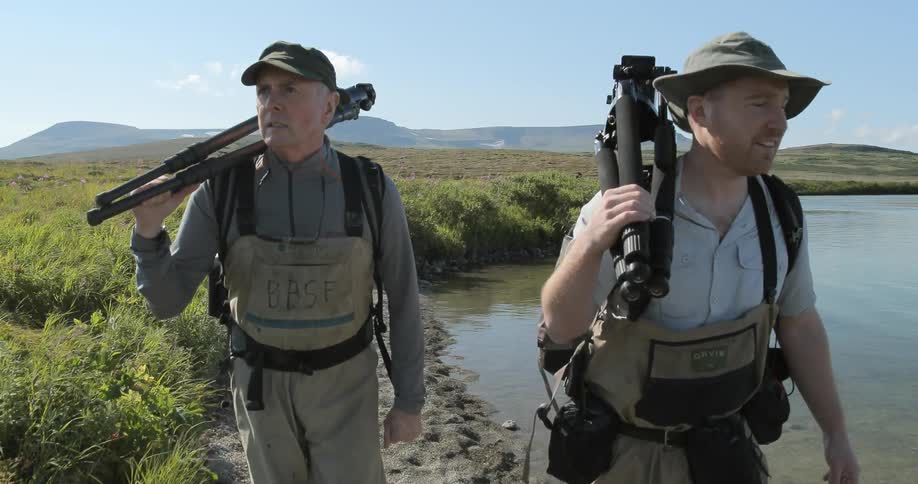 Two people carrying tripods are walking near a stream on a sunny day