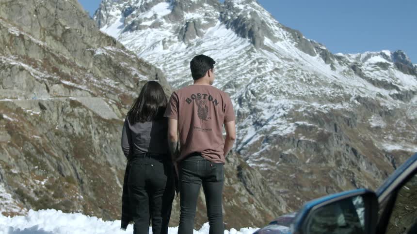 A man and woman stand in the snow next to their car gazing at the mountains