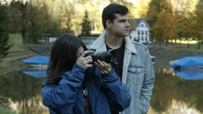 A man and a woman with a camera stand by a lake looking into the distance
