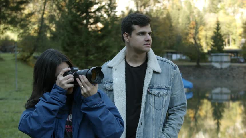 Two people stand next to a lake while one person takes a picture with a camera