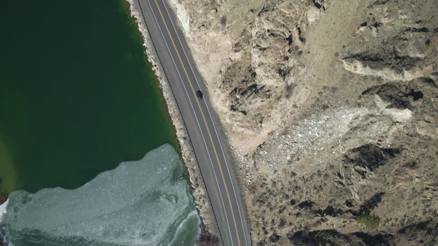 Birds eye view of a car driving fast on a highway between a rugged mountainside and partially frozen lake