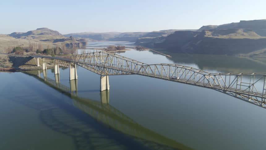 A car drives on a long bridge over calm water with tall canyon cliffs in the distance