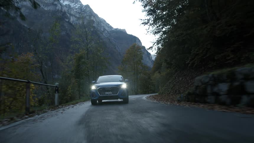 A blue car travels down a road next to a forest and mountain valley
