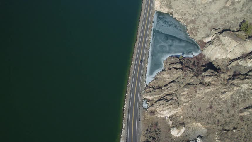 A car drives on a road between a deep cold lake and mountainside with a frozen pond