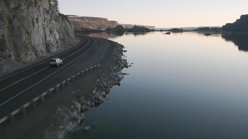 Fast chase behind a silver SUV as it drives down a road next to a perfectly calm lake surrounded by rocky cliffs
