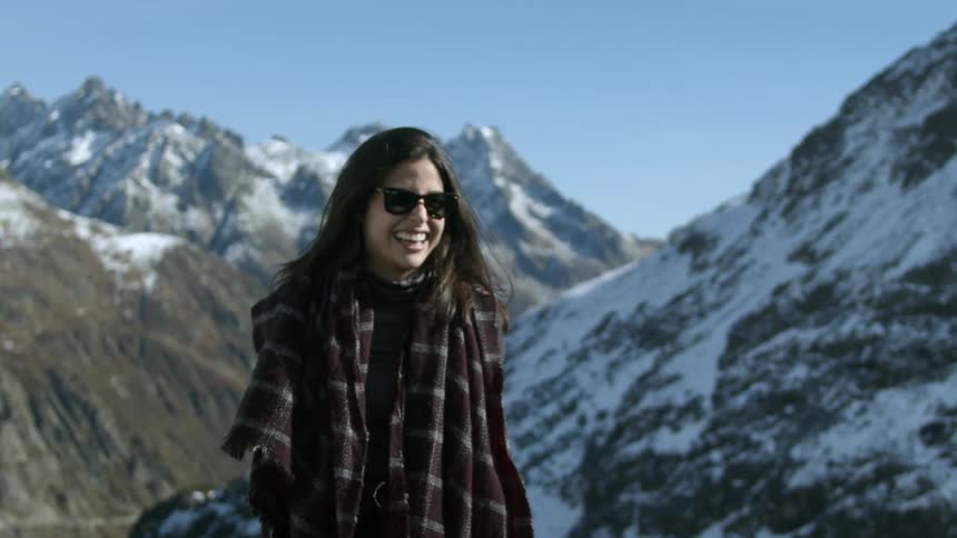A woman wearing a scarf and sunglasses smiles big as she walks with mountains behind her