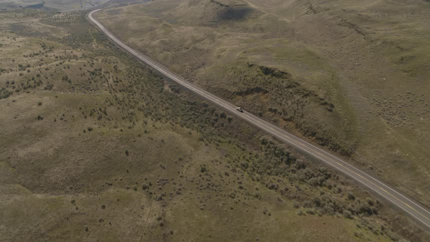 Wide overview of a pickup truck driving on a long stretch of winding road through green foothills