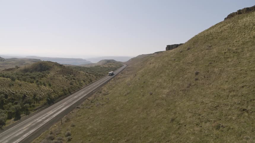 A large semi truck hauls a load on a rural highway through rugged green foothills on a sunny day