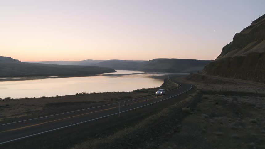 Intersecting a white car driving on an S curve beside a mountain lake at dawn